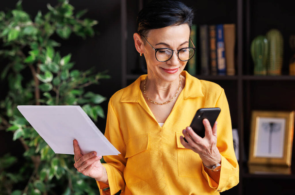 Reife Managerinnen in stylischer gelber Bluse und Brille lesen während der Arbeit im Home Office Daten auf Papier und surfen in den sozialen Medien auf dem Kundenhandy - Foto, Bild
