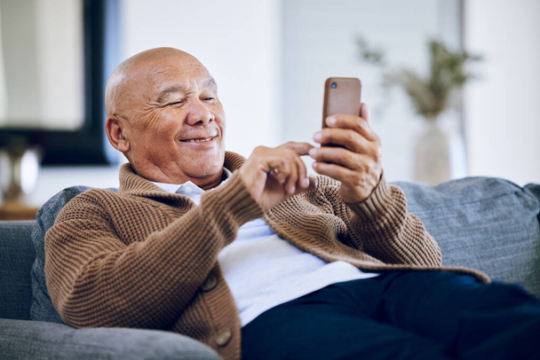 Mosolyogj, gépelj, és egy idős ember telefonnal a kanapén a közösségi média, a kapcsolat vagy a vesszőzés. Happy, pihenjen és egy idős személy a kanapén egy mobil alkalmazás vagy olvasás egy chat. - Fotó, kép