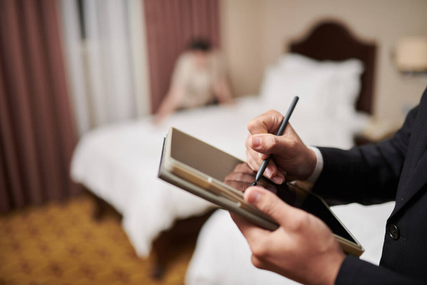 Lähikuva kuva hotellipäällikkö täyttö lomake välilehti anna tietokoneen kun piika petaa sängyn taustalla - Valokuva, kuva
