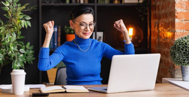 Szczęśliwy w średnim wieku kobieta biznesu w modnym niebieskim golfie i okulary podnoszące ręce i patrząc na ekranie laptopa z uśmiechem po zakończeniu projektu siedząc w biurze w domu - Zdjęcie, obraz