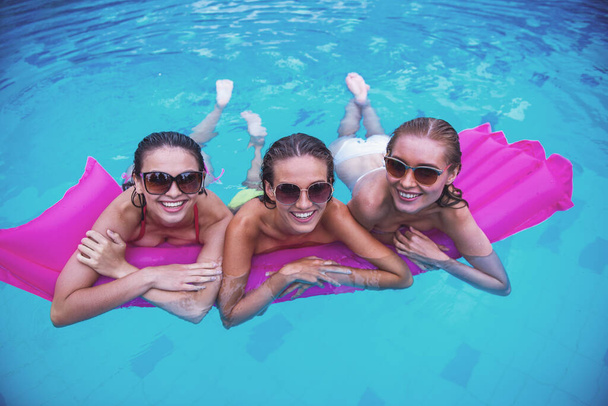 Kauniit tytöt uimapuvuissa ja aurinkolaseissa katsovat kameraa ja hymyilevät uima-altaassa
 - Valokuva, kuva