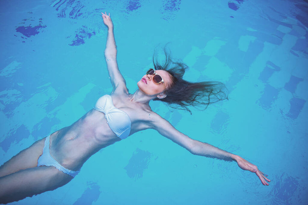 Высокий угол зрения привлекательной чувственной девушки в белых купальниках и солнцезащитных очках, плавающих на спине в бассейне
 - Фото, изображение