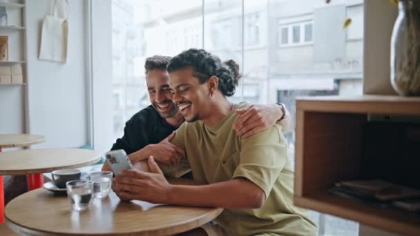 Homosexual family video calling sitting small restaurant at romantic date close up. Happy multiracial gay partners greeting to phone camera. Smiling men lovers enjoy online communication in cafeteria. - Footage, Video
