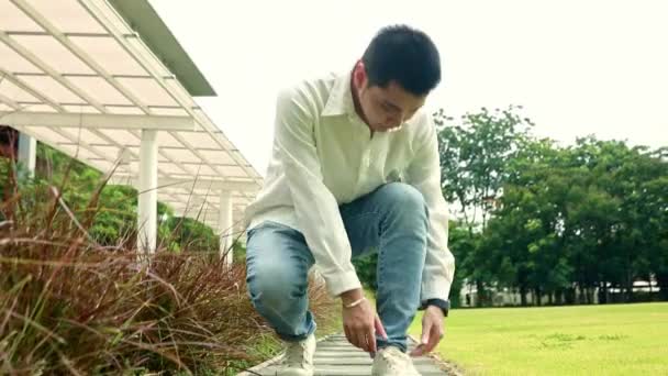 Jeune homme asiatique beau arrête de marcher pour s'asseoir et attacher ses lacets lâches dans l'herbe et regarder la caméra de bonne humeur. - Séquence, vidéo