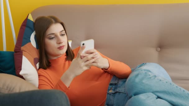 Сконцентрированная девушка, печатающая на смартфоне дома. Молодая женщина лежит на диване. Улыбающаяся женщина с мобильного телефона. Студентка отдыхает после онлайн-уроков. Веселая модель просмотра интернета на мобильном телефоне - Кадры, видео