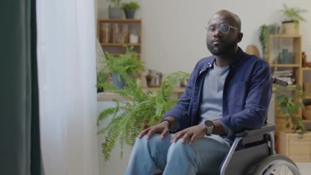 Retrato de hombre afroamericano con discapacidad sentado en silla de ruedas y posando para la cámara al lado de la ventana en casa - Metraje, vídeo