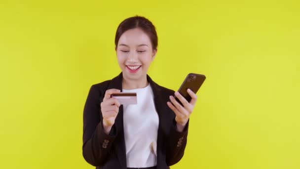 Portrait young asian business woman hold credit card and looking smart phone for shopping online isolated yellow background, businesswoman looking debit card and smartphone, financial concept. - Footage, Video