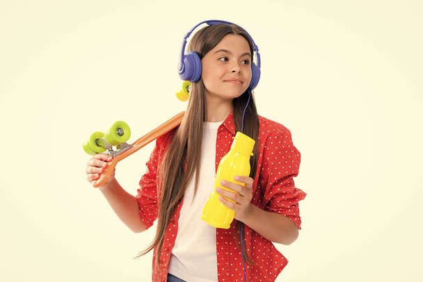 Teen girl 12, 13, 14 years old with skateboard water bottle and headphones over studio background. Cool modern teenager in stylish clothes. Teenagers lifestyle, casual youth culture - Foto, Bild