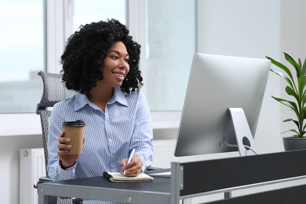 Young woman with cup of drink taking notes near computer at table in office - Foto, Bild