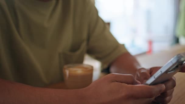 Muž ruce drží smartphone na dřevěném stolku kavárny zblízka. Mladý latinskoamerický muž vypadá mobilní telefon obrazovka sedí kavárna sám. Tmavé vlasy hipster zprávy na mobilu v útulné kavárně. - Záběry, video
