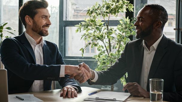 Two successful diverse businessmen sit at office desk with laptop happy shake hands. Caucasian man salesman manager handshake satisfied African American client customer investor at business meeting - 写真・画像