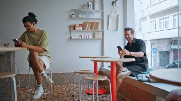 カフェテリアに座っている2人の男がスマートフォンの画面を見ています. 快適なコーヒーハウスのインテリアで個別にリラックスしたリラックスしたマルチエスニックカフェ. 携帯電話でアロマティックコーヒーを飲むカップル男性 - 映像、動画