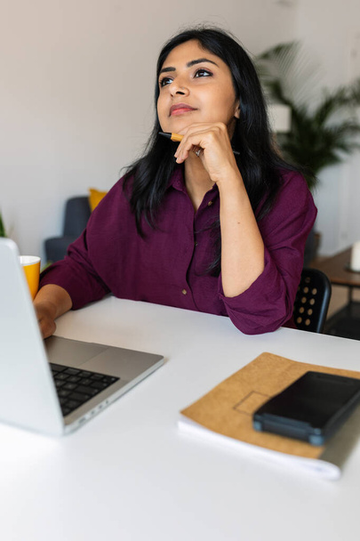 Ragionevole donna indiana seduta con la mano sul mento utilizzando il computer portatile sul tavolo mentre distoglie lo sguardo, pensando a idee di business o problemi personali. - Foto, immagini
