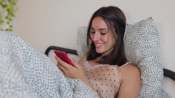Joven niña milenaria divirtiéndose usando el teléfono celular descansando en la cama. Millennial mujer leyendo mensaje divertido en la aplicación del teléfono inteligente después de despertar. - Metraje, vídeo