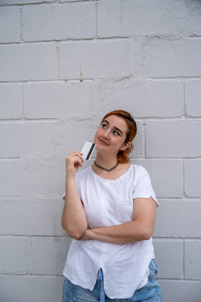 Jeune femme hispanique positive tenant une carte de crédit avec bande magnétique dans les doigts près du visage et levant les yeux tout en se tenant avec la main croisée près du mur de briques grises en plein jour - Photo, image