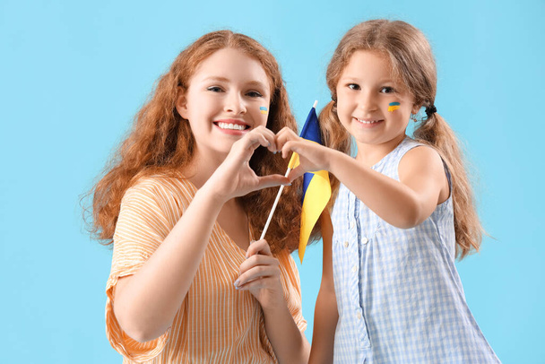 Счастливая маленькая девочка и ее мать с украинским флагом показывают жест сердца на синем фоне - Фото, изображение