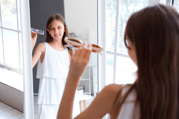 Stijlvol klein meisje met nieuwe zonnebril kijken in spiegel thuis - Foto, afbeelding