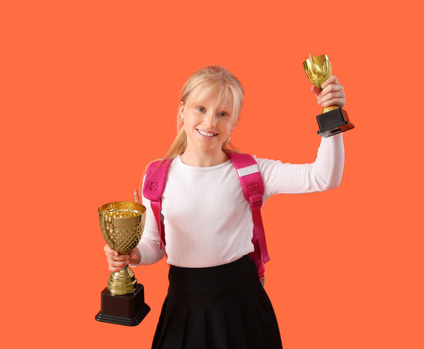 Умная маленькая школьница в стильной форме с призовыми кубками на оранжевом фоне - Фото, изображение