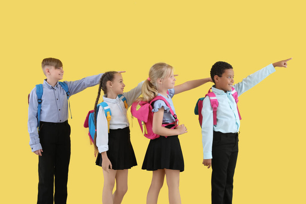 Kleine Klassenkameraden in stylischer Uniform mit Rucksäcken, die auf etwas auf gelbem Hintergrund zeigen - Foto, Bild