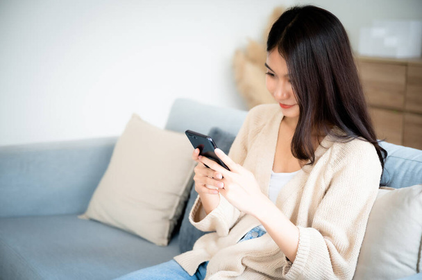 Happy young asian woman relax on comfortable couch at home texting messaging on smartphone, smiling girl use cellphone chatting, browse wireless internet on gadget, shopping online from home - Photo, Image