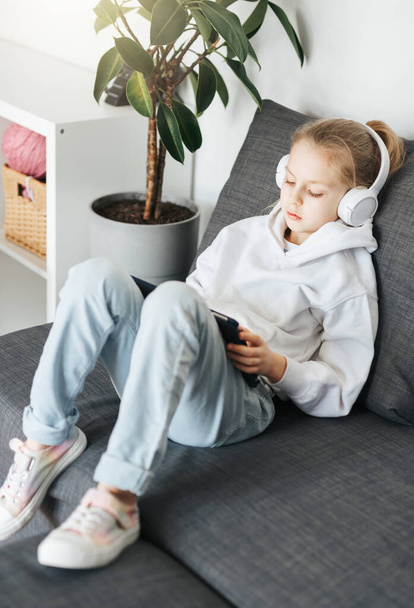 A kislány, aki a kanapén fekszik egy elektronikus táblagéppel, zenét hallgat fehér fülhallgatóban. Vezeték nélküli eszközök használata az oktatásban. - Fotó, kép
