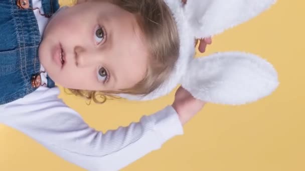 Een schattig klein meisje met paashaas oren op een gele achtergrond toont verschillende emoties, vreugde, verrassing, geluk, dromen. Verticale video - Video