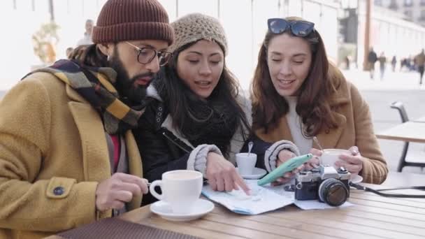 Туристичні друзі дивляться на смартфон, сидячи в кафе, перевіряючи карту міста під час зимових канікул  - Кадри, відео