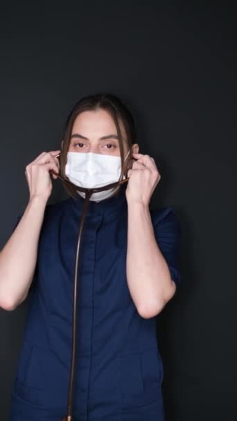 Imagem vertical. Retrato de uma jovem médica em um fundo escuro. Um médico em uma máscara médica coloca um tubo, um estetoscópio. Médico-terapeuta. Pediatra. Vídeo vertical - Filmagem, Vídeo