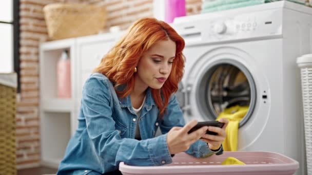 Młoda ruda kobieta za pomocą smartfona pranie ubrania w pralni - Materiał filmowy, wideo