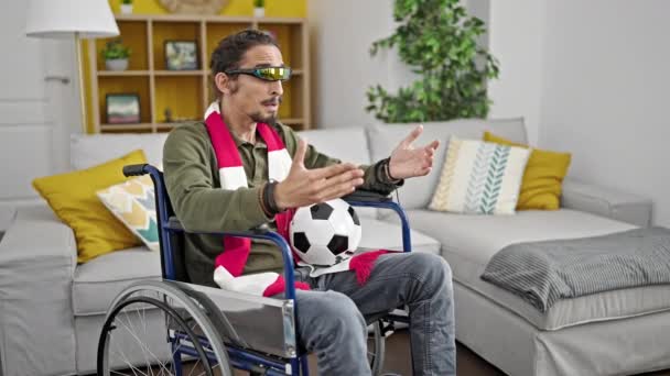 Jovem hispânico assistindo jogo de futebol usando óculos de realidade virtual sentado em cadeira de rodas em casa - Filmagem, Vídeo