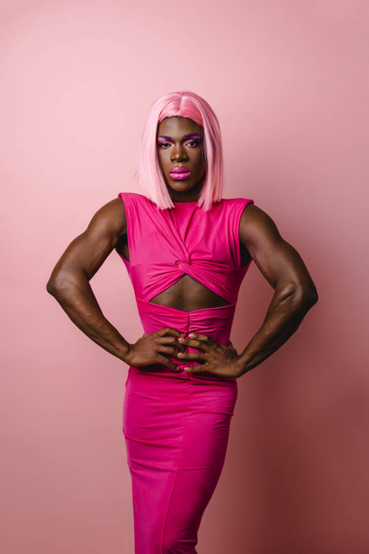 Уверенный африканский трансгендер в розовом платье и парике, смотрящий в камеру, позируя на изолированном розовом фоне. - Фото, изображение