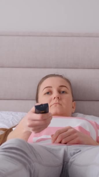 A woman watches TV, cant fall asleep, doesnt sleep well. A young woman is lying on a bed with a TV remote control in her hands. Depressed woman spends time alone. Vertical video - Footage, Video