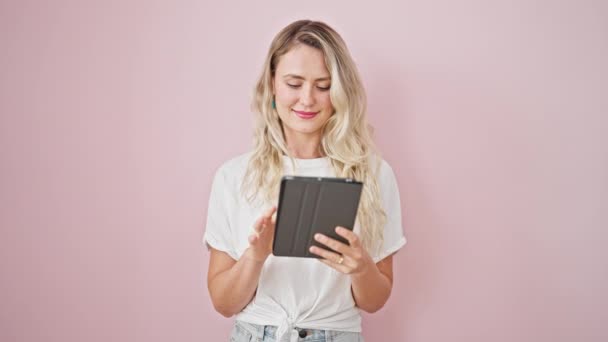 Giovane donna bionda sorridente fiducioso utilizzando touchpad su isolato sfondo rosa - Filmati, video