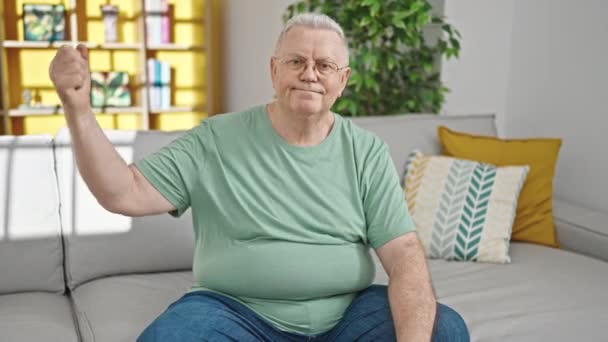 Středního věku šedovlasý muž usměvavý sebevědomě sedí na pohovce dělá silné gesto doma - Záběry, video