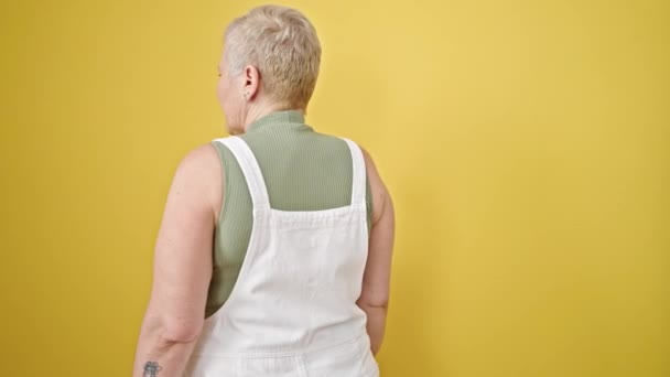 Mujer de pelo gris de mediana edad sonriendo confiada de pie sobre un fondo amarillo aislado - Metraje, vídeo