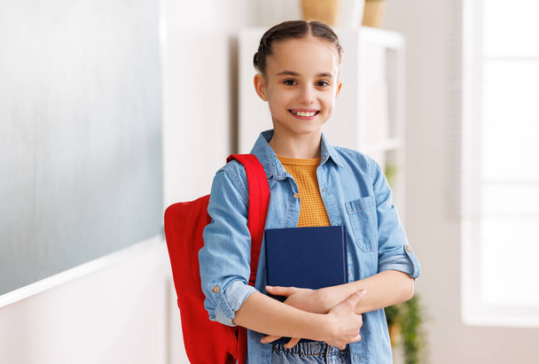 Positiv cleveres Mädchen in lässiger Kleidung mit Rucksack und Zöpfen, lächelnd und in die Kamera blickend, während es in einem sonnendurchfluteten Klassenzimmer in der Schule steht - Foto, Bild