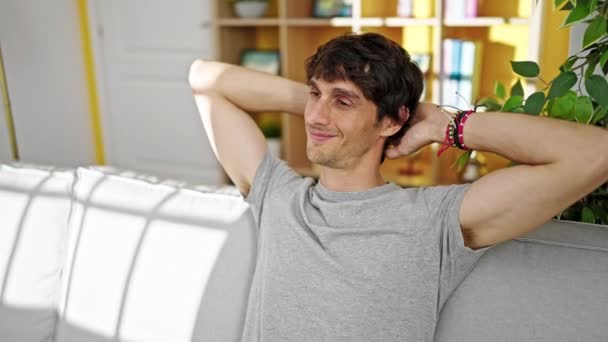 Jonge Spaanse man ontspannen met handen op het hoofd zittend op de bank thuis - Video