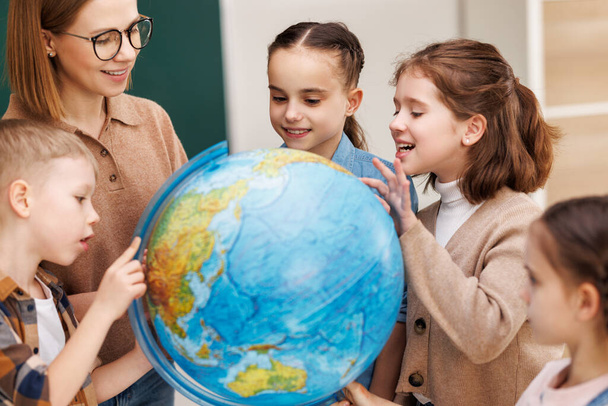 Donna in occhiali e bambini positivi esaminando globo mentre in piedi intorno al tavolo in classe di geografia durante la lezione a scuola - Foto, immagini