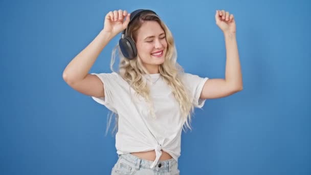 Junge blonde Frau hört Musik und tanzt vor isoliertem blauen Hintergrund - Filmmaterial, Video