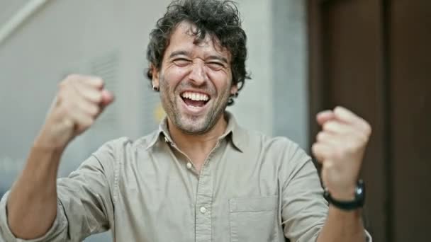 Jonge Spaanse man glimlachend vol vertrouwen staand met winnaar gebaar op straat - Video