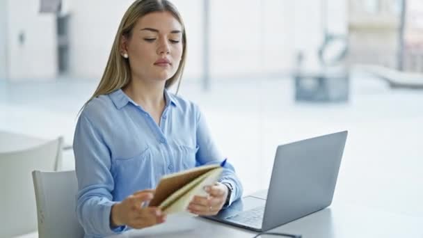 Νεαρή ξανθιά εργαζόμενη επιχείρηση χρησιμοποιώντας φορητό υπολογιστή ανάγνωση σημειωματάριο στο γραφείο - Πλάνα, βίντεο