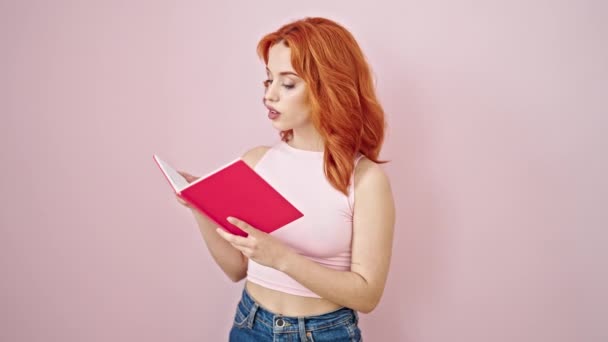 Giovane rossa donna lettura libro facendo pollice verso l'alto gesto su isolato sfondo rosa - Filmati, video