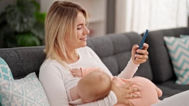 Madre e hija sentadas en el sofá amamantando al bebé usando smartphone en casa - Metraje, vídeo