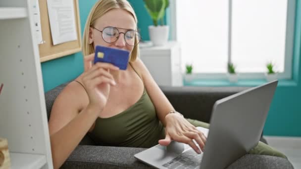 Joven mujer rubia trabajadora de negocios de compras con laptop y tarjeta de crédito celebrando en la oficina - Imágenes, Vídeo