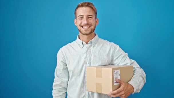 Young caucasian man holding package doing thumb up gesture over isolated blue background - Footage, Video