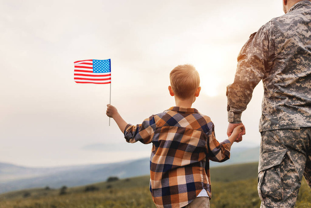 Hátsó kilátás katonai ember apa fogja fia kezét amerikai zászló és élvezi a csodálatos nyári természet kilátás napsütéses napon, boldog férfi katona apa újra együtt fia után amerikai hadsereg - Fotó, kép