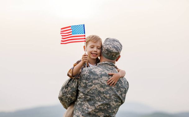 Happy little boy son with American flag hugging father in military uniform came back from US army, rear view of male soldier reunited with family while standing in green meadow on summer day - Photo, Image