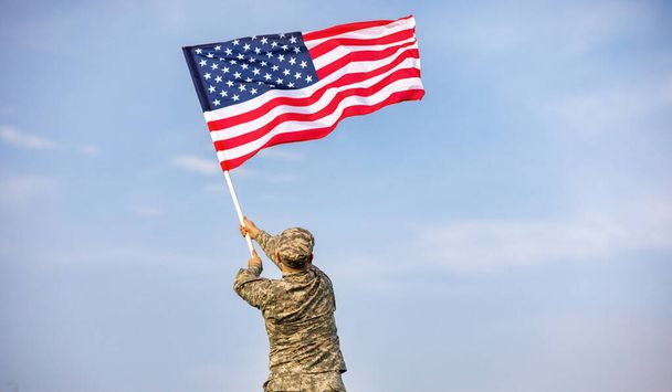 Vista dal retro di un soldato di sesso maschile in uniforme dell'esercito americano che sventola la bandiera degli Stati Uniti sulla cima di una montagna in una radura al tramonto - Foto, immagini