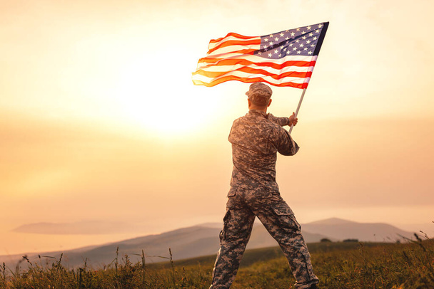 Άποψη από το πίσω μέρος ενός άνδρα στρατιώτη με τη στολή του αμερικανικού στρατού κουνώντας τη σημαία των ΗΠΑ στην κορυφή ενός βουνού σε ένα ξέφωτο το ηλιοβασίλεμα - Φωτογραφία, εικόνα
