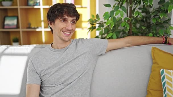 Fiatal spanyol férfi néz oldalra ül a kanapén és otthon mosolyog. - Felvétel, videó